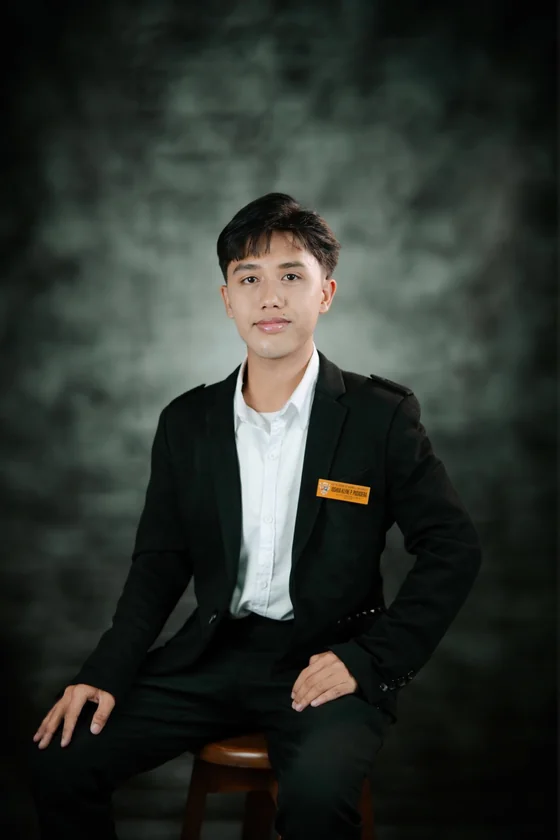Studio portrait of Joshua Klyne Pudadera seated on a wooden stool in a black blazer over an open white dress shirt, a Notre Dame of Marbel University name badge pinned to his jacket, against a dark textured backdrop